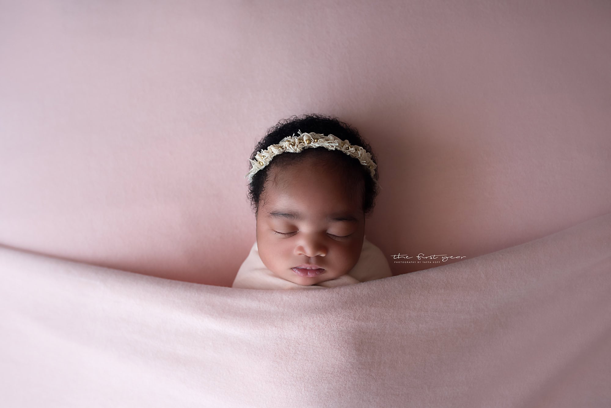 Adorable sleeping newborn girl with floral headband, captured in a soft pink setting.