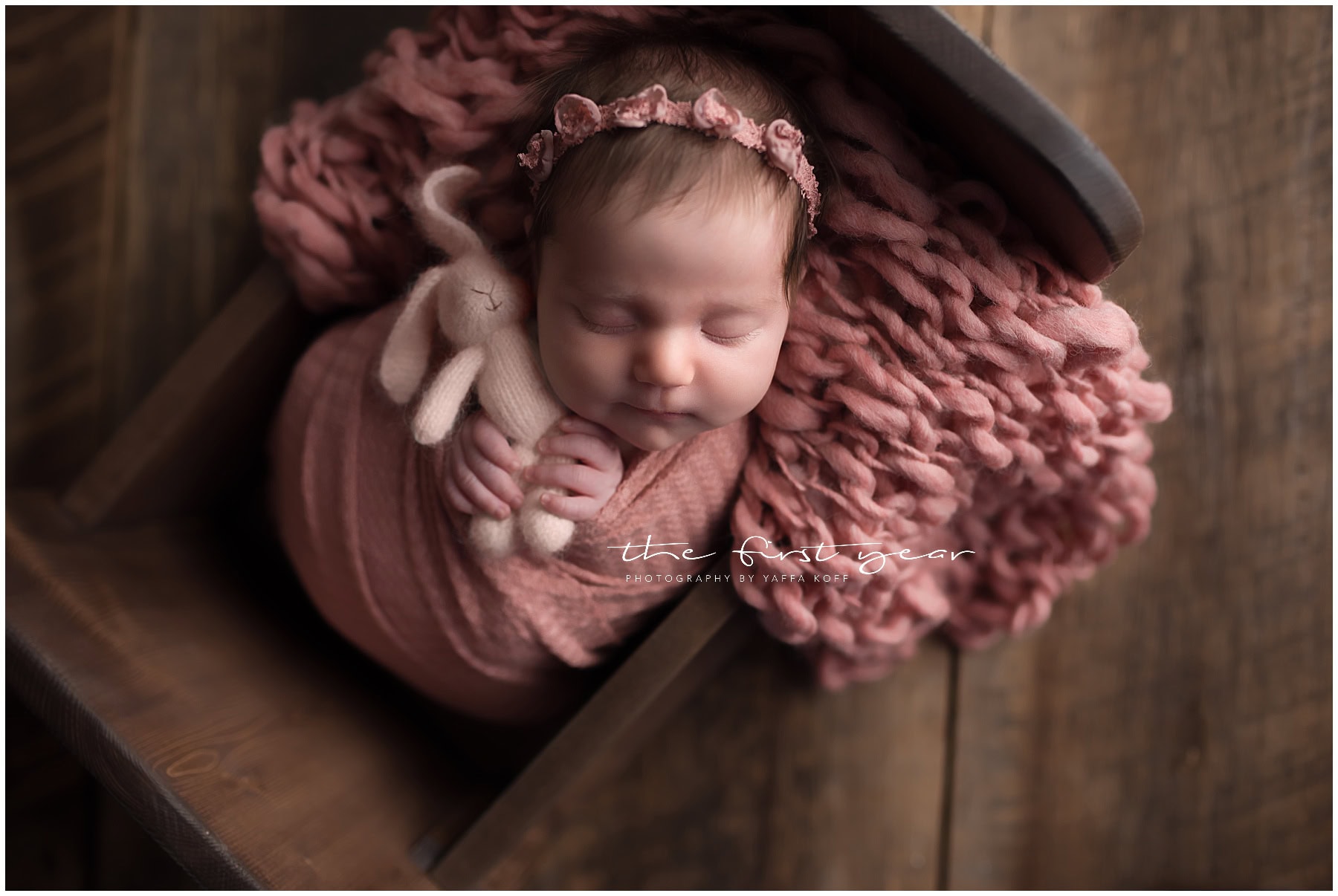 Baby sleeping peacefully in a cozy wooden crate with soft pink blanket and headband.