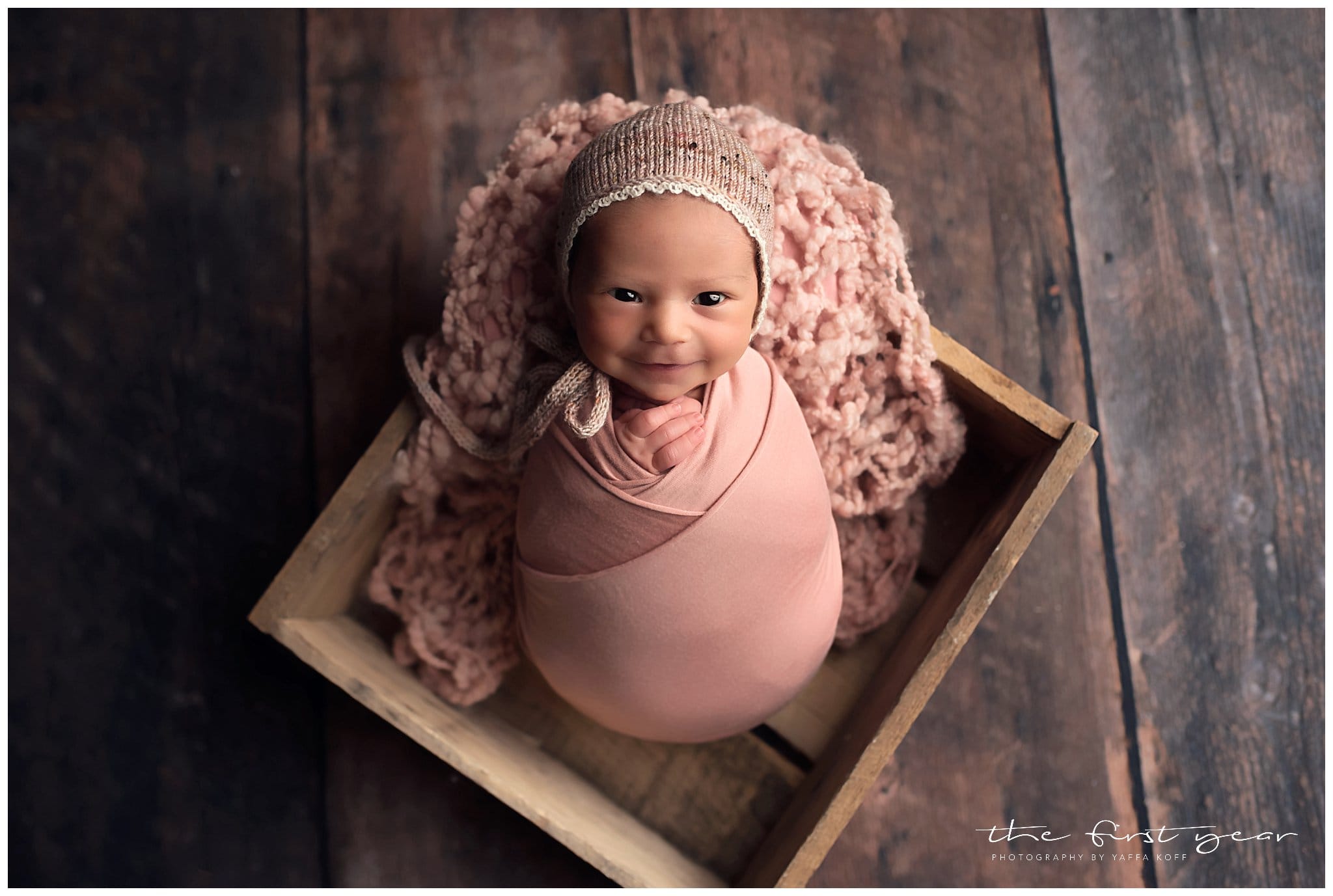 Baby girl wrapped in pink with a cozy knit hat, sitting in a wooden crate on a dark wooden floor.