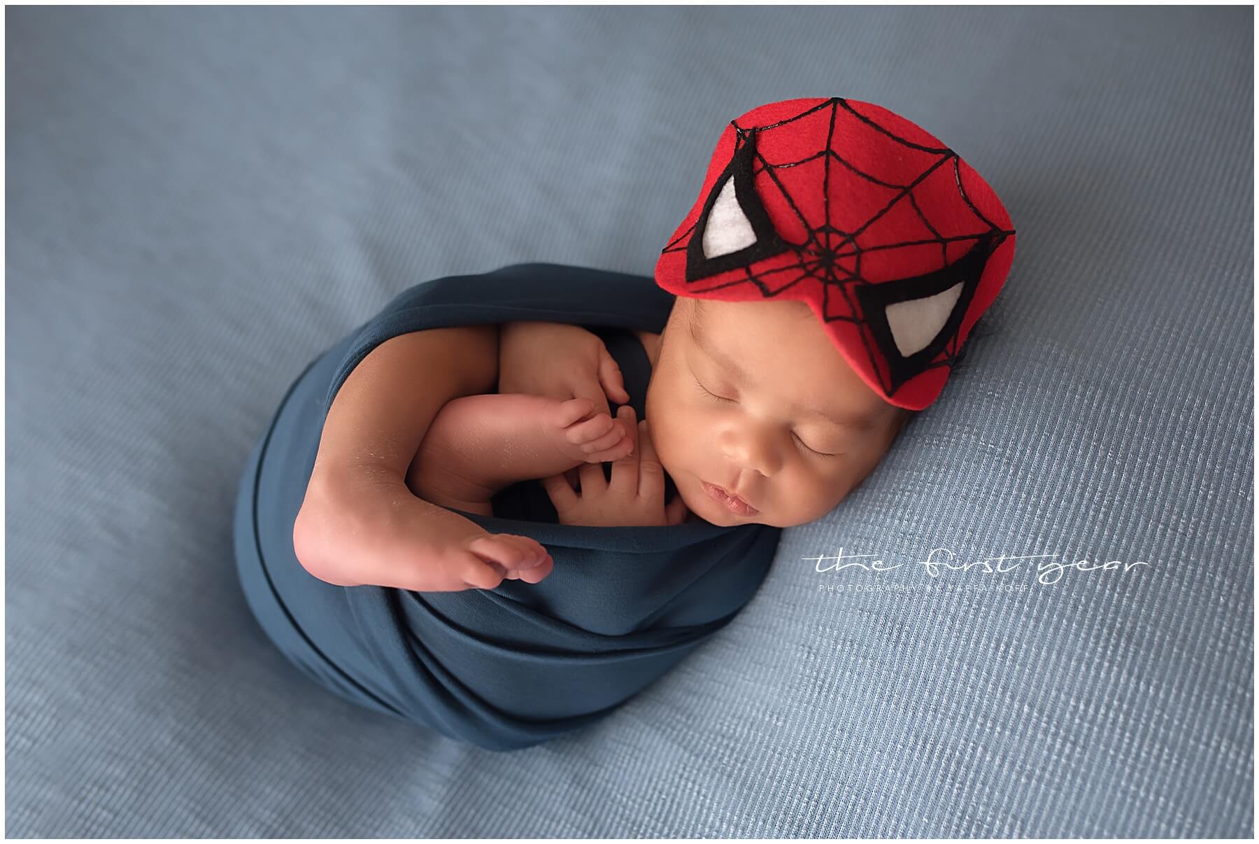 Newborn baby sleeping peacefully wearing a Spiderman hat, wrapped in a soft blue blanket.