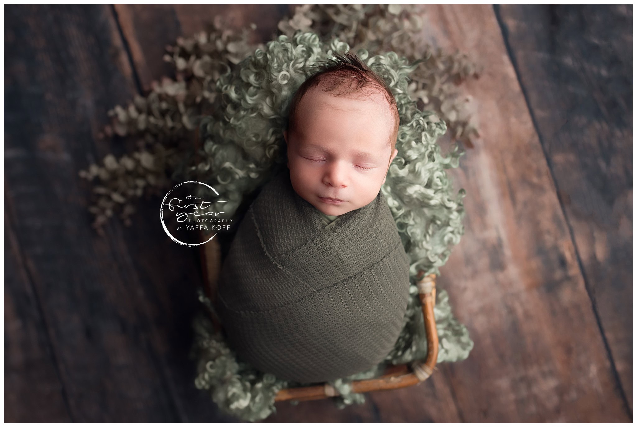 Adorable newborn baby wrapped in green, sleeping peacefully in a basket with greenery backdrop.
