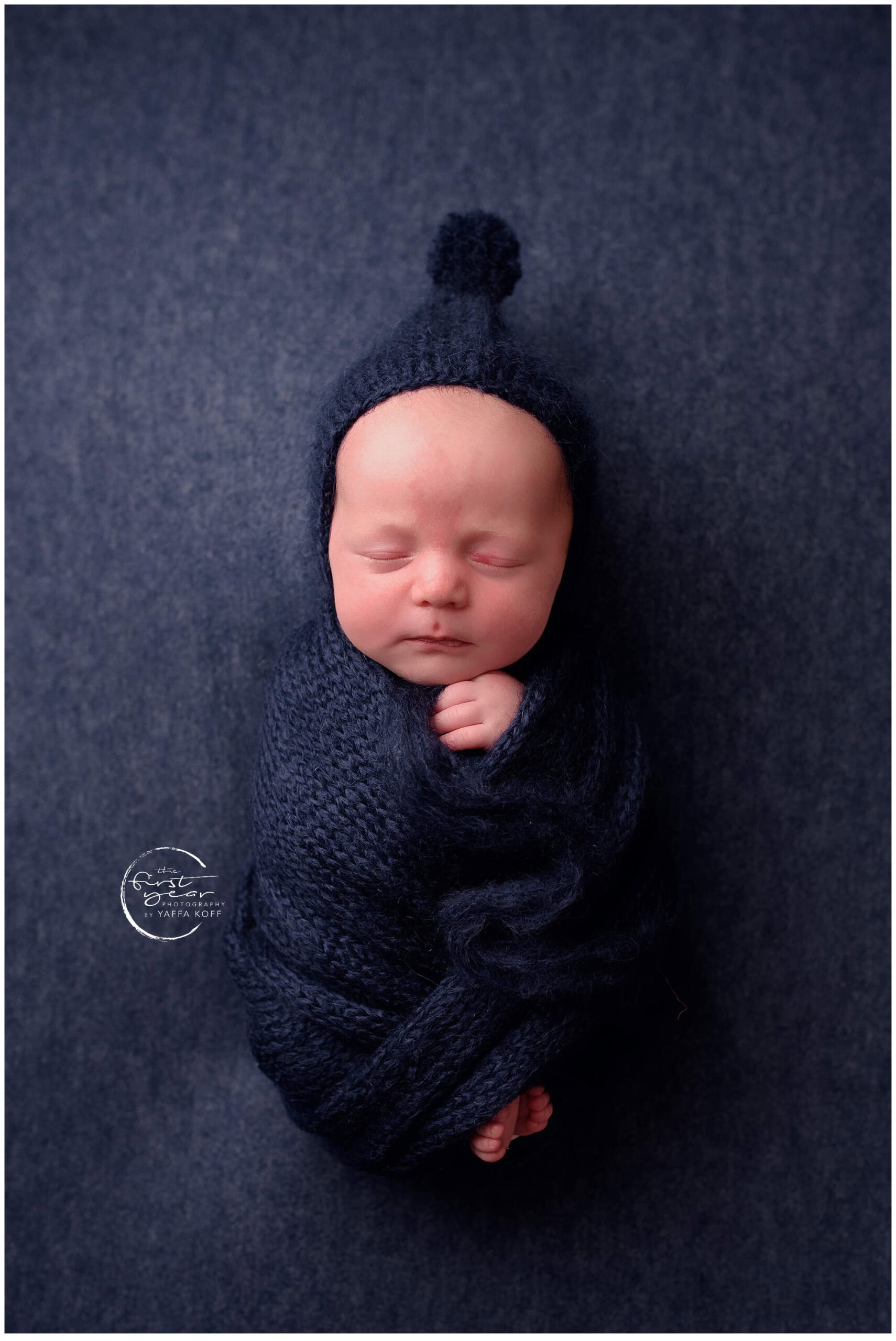 Newborn baby Yehuda wrapped in navy blue with matching hat, peacefully sleeping.