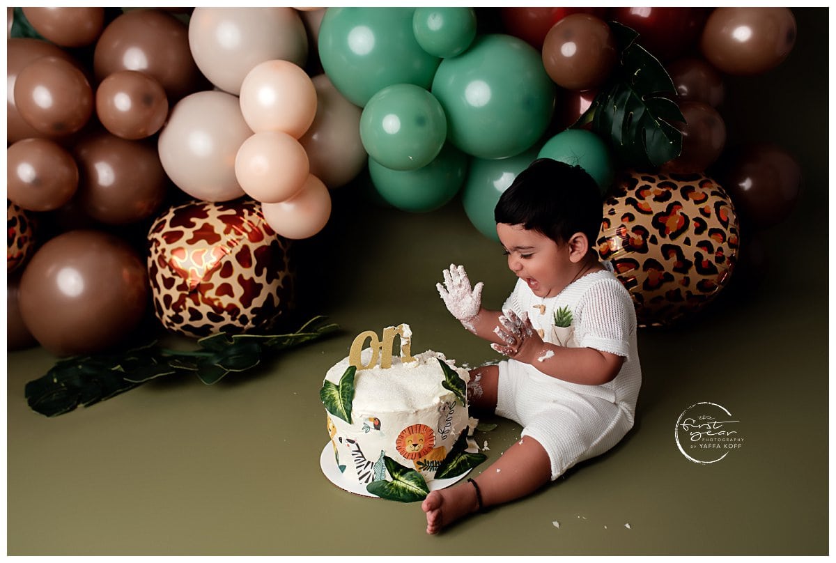 Child enjoying his first birthday cake with colorful balloons backdrop.