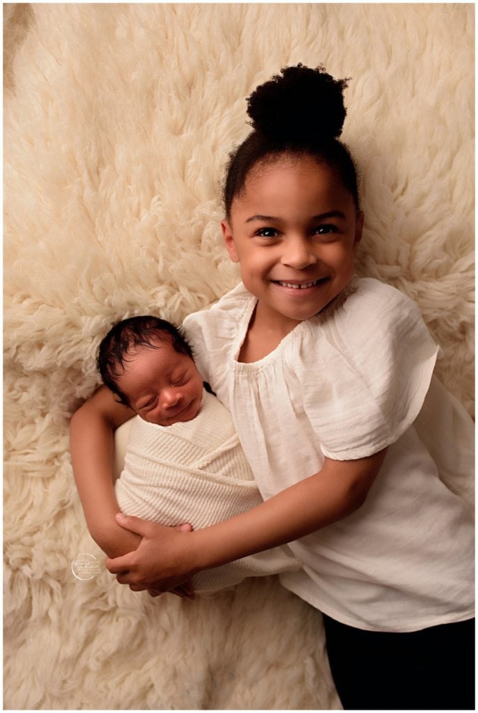 Adorable newborn with older sister lying on fluffy cream blanket.