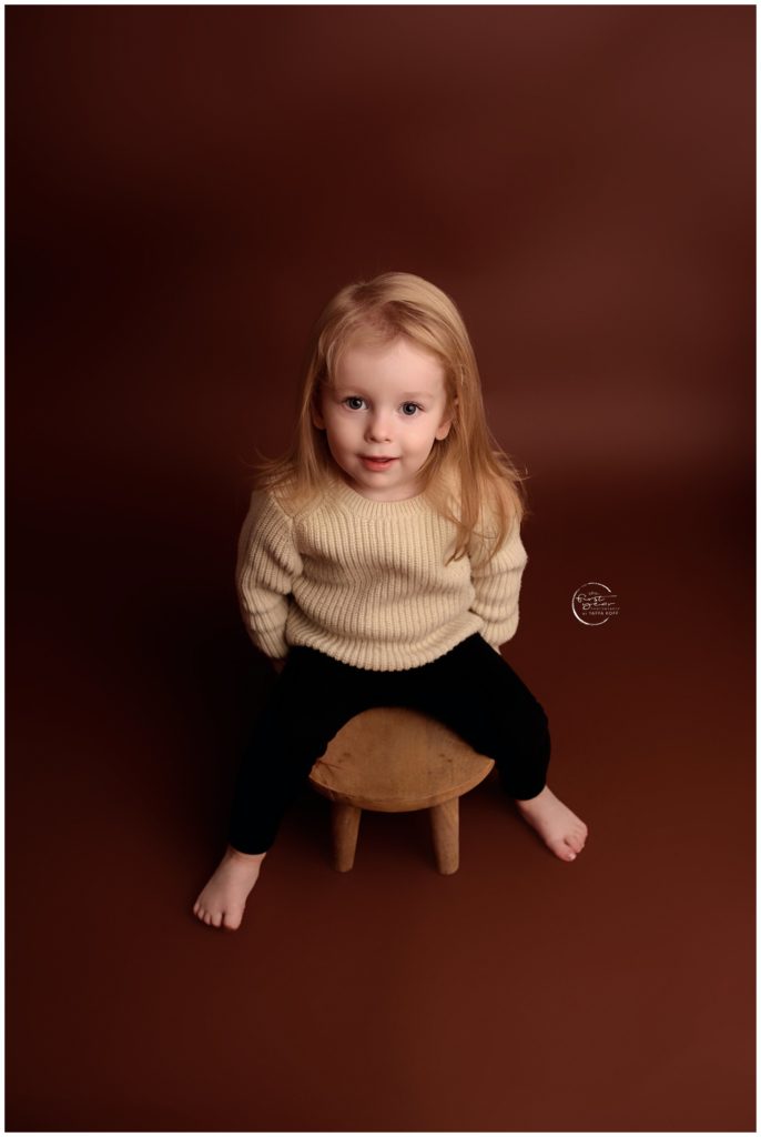 Adorable young girl with blonde hair sitting on a small stool against a brown background.