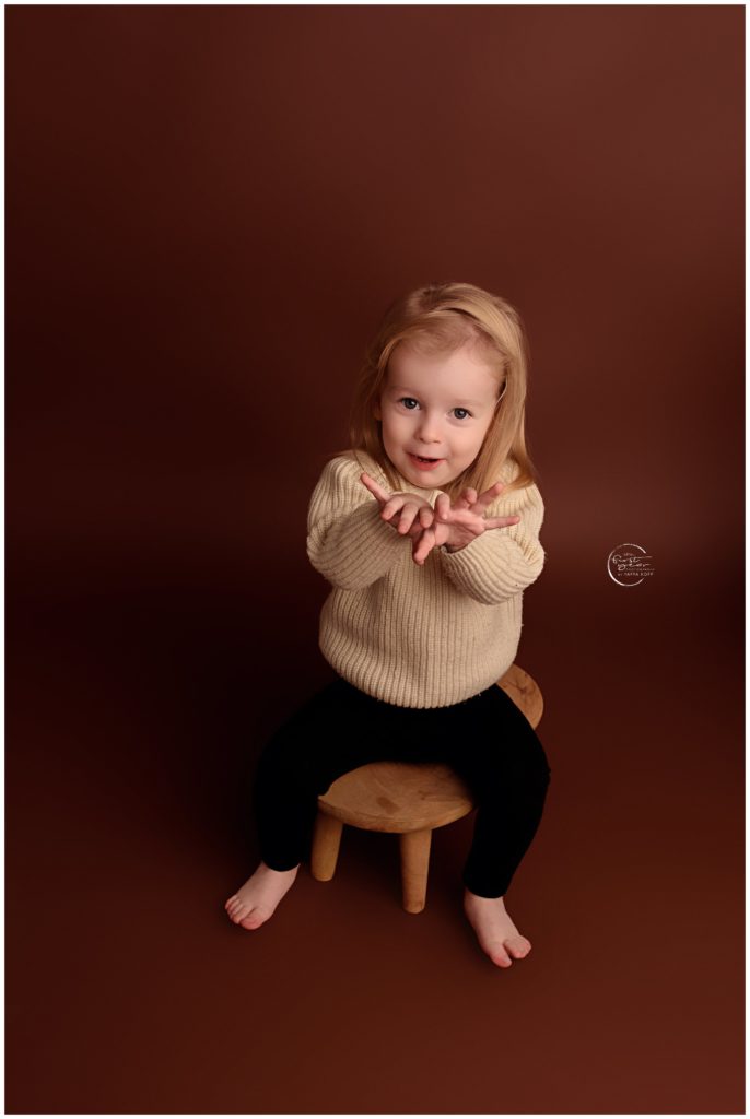 Adorable young girl sitting on a stool against a brown background.