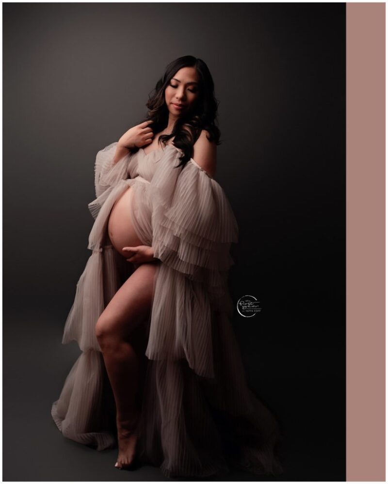 Pregnant woman in flowing gown holding belly, studio maternity photo.