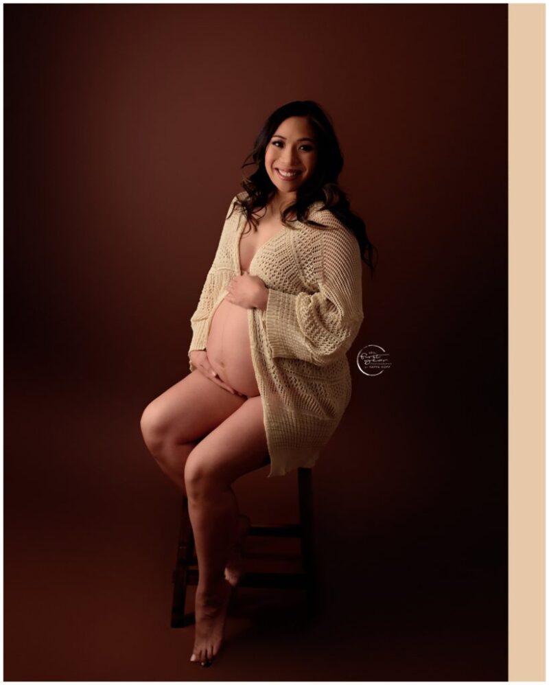 Pregnant woman sitting on a stool, smiling, in a maternity photoshoot.