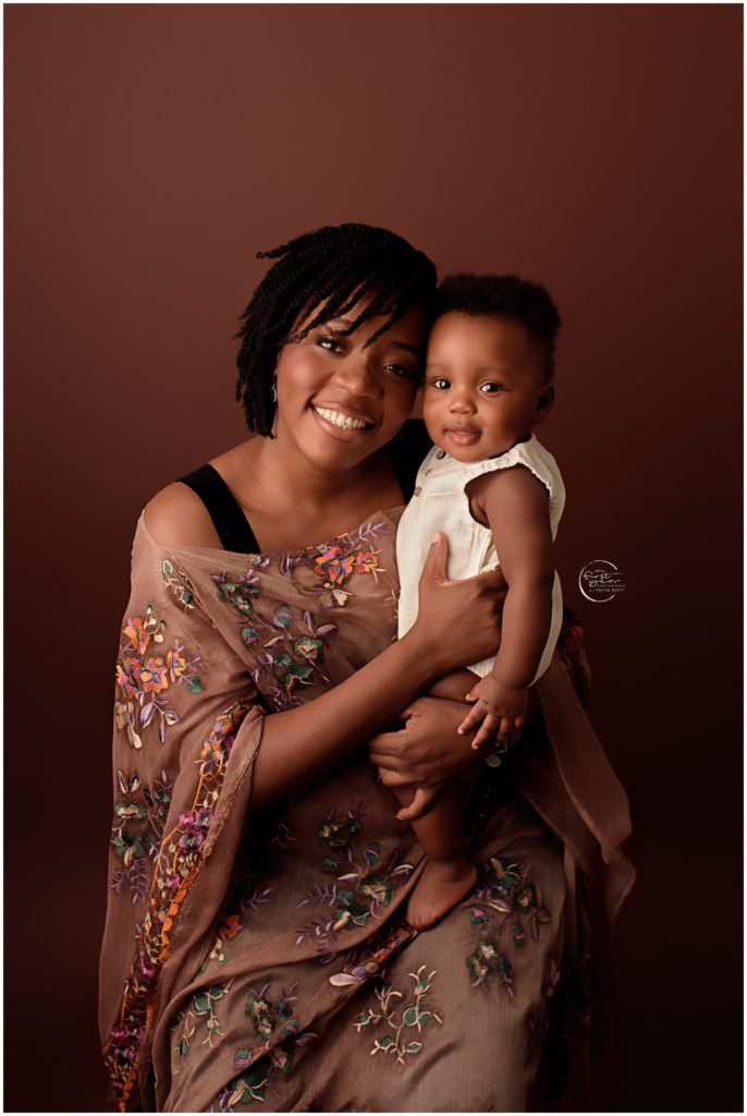 Mother holding her smiling baby girl against a warm brown background.