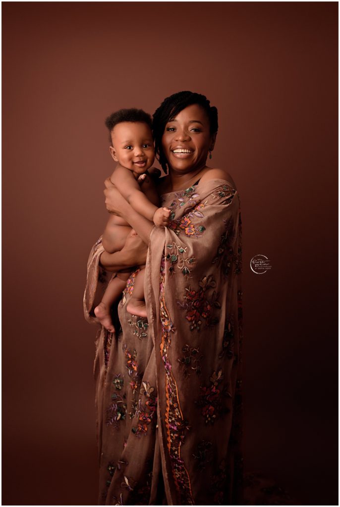 Mother holding her smiling baby girl, both dressed elegantly against a warm brown background.