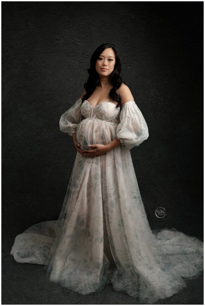 Pregnant woman in flowing gown holding belly, studio maternity photo.