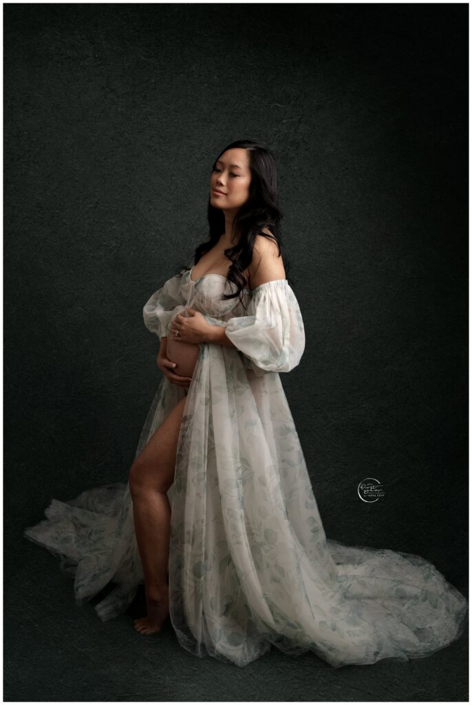 Pregnant woman in flowing lace gown, gently holding her belly, against a dark textured background.