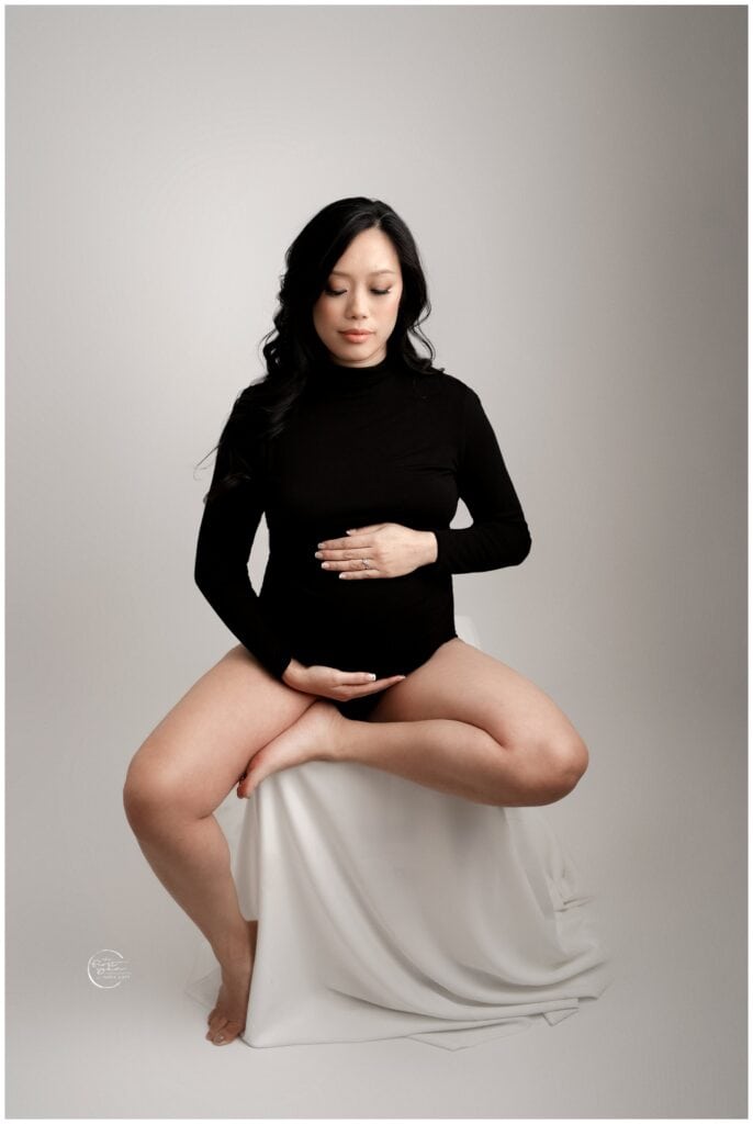 Pregnant woman sitting cross-legged on a white stool, gently holding her belly, in a professional ma.