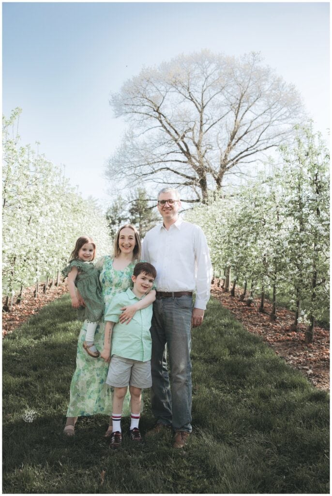 Family portrait of four standing on a grassy path among blossoming trees during spring.