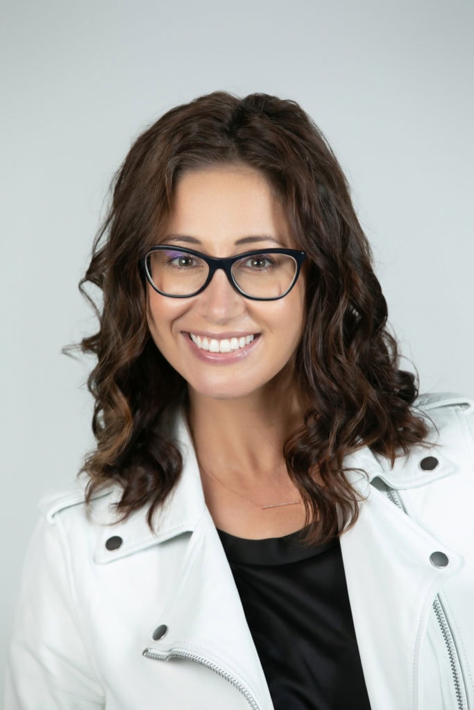 Portrait of a smiling woman with glasses, white jacket, and curly hair.